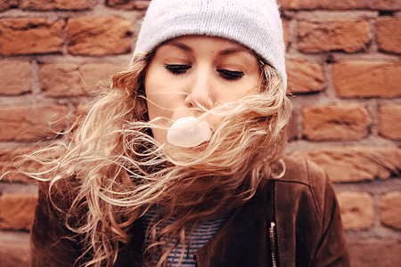 Teen girl blowing a pink bubble gum bubble with a brick wall behind her.