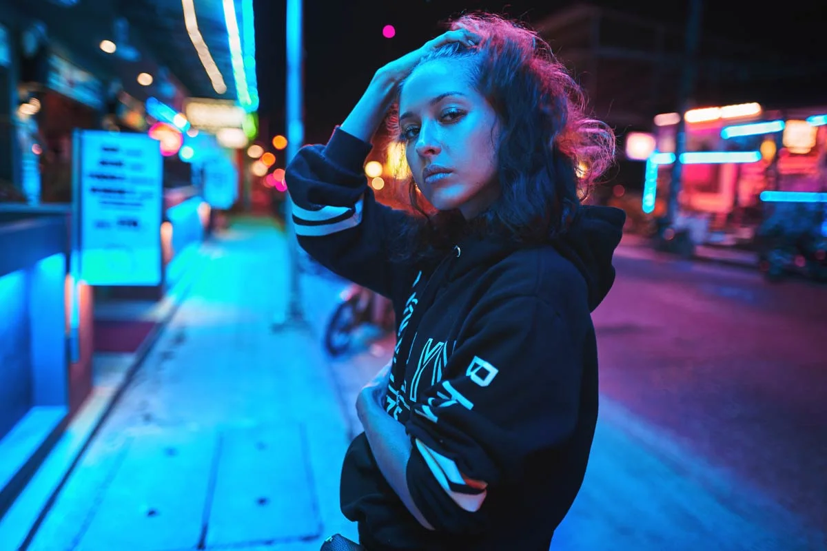 A woman in a hoodie stands on a street at night. Neon lights in blue and pink illuminate the scene. A woman in a hoodie stands on a street at night. Neon lights in blue and pink illuminate the scene.