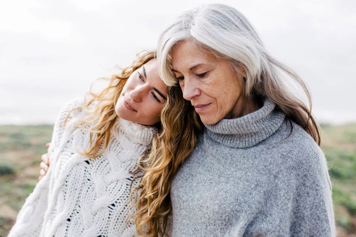 Due donne con i capelli lunghi sono in piedi all'esterno, la più giovane appoggia la testa alla spalla della più anziana. Due donne con i capelli lunghi sono in piedi all'esterno, la più giovane appoggia la testa alla spalla della più anziana.