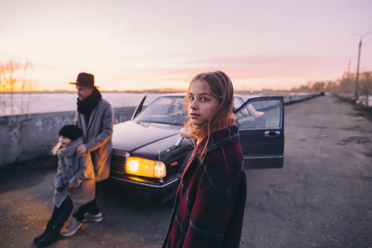 Person in front of a car with light, two people in the background, river and sunset visible. Person in front of a car with light, two people in the background, river and sunset visible.