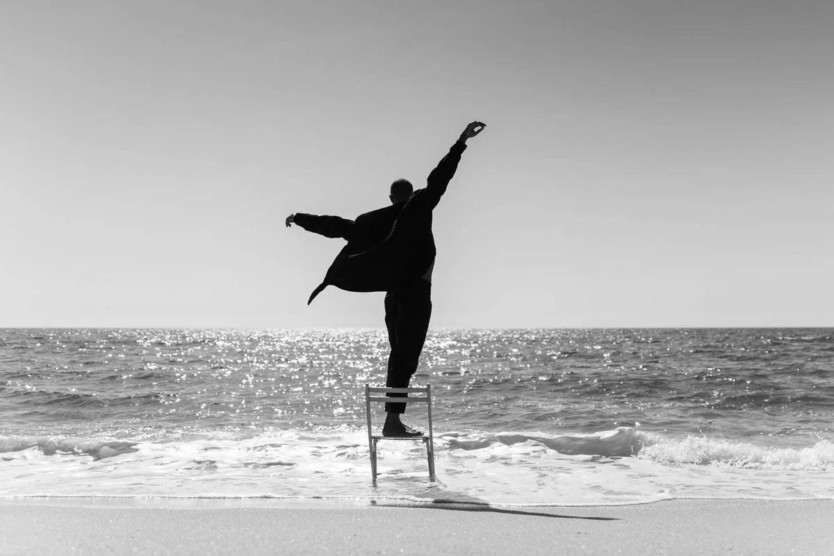 Person on a chair by the sea, arms outstretched. Black and white shot, water glistening in the background. Person on a chair by the sea, arms outstretched. Black and white shot, water glistening in the background.