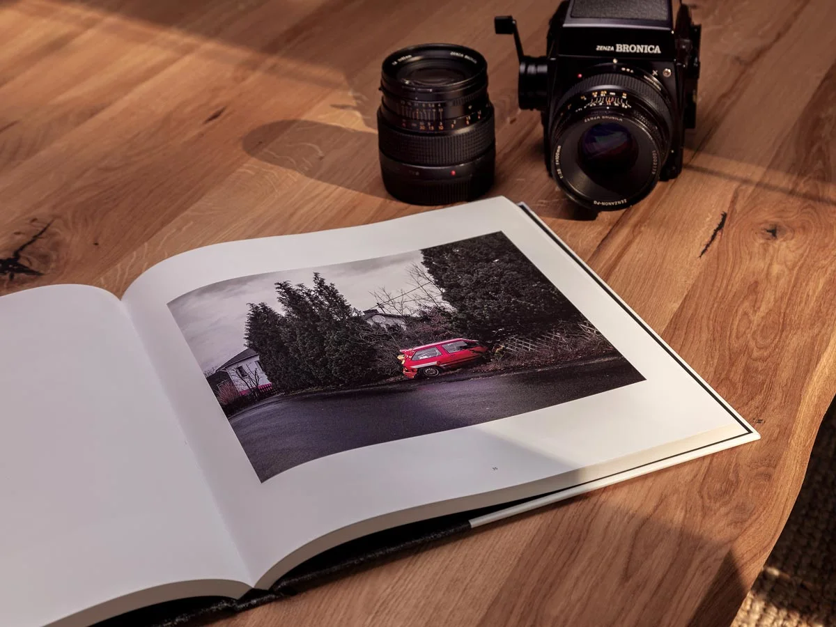 Ein Bildband liegt auf einem Holztisch, aufgeschlagen mit einem Foto eines roten Autos vor Büschen. Ein Bildband liegt auf einem Holztisch, aufgeschlagen mit einem Foto eines roten Autos vor Büschen.