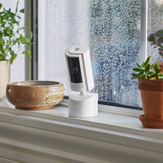 White Ring Pan-Tilt Indoor Cam sits on windowsill with rainy window in the background.