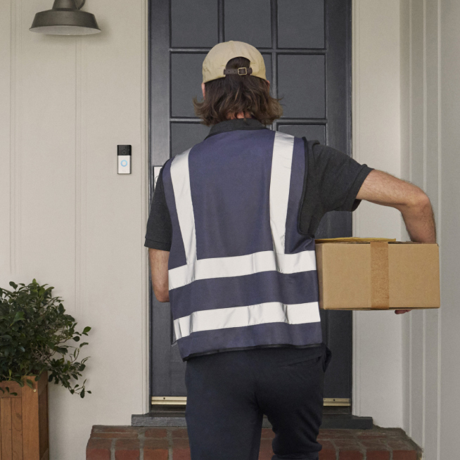 A delivery person holding a package approaches a black front door, with a Ring Battery Doorbell mounted beside it.