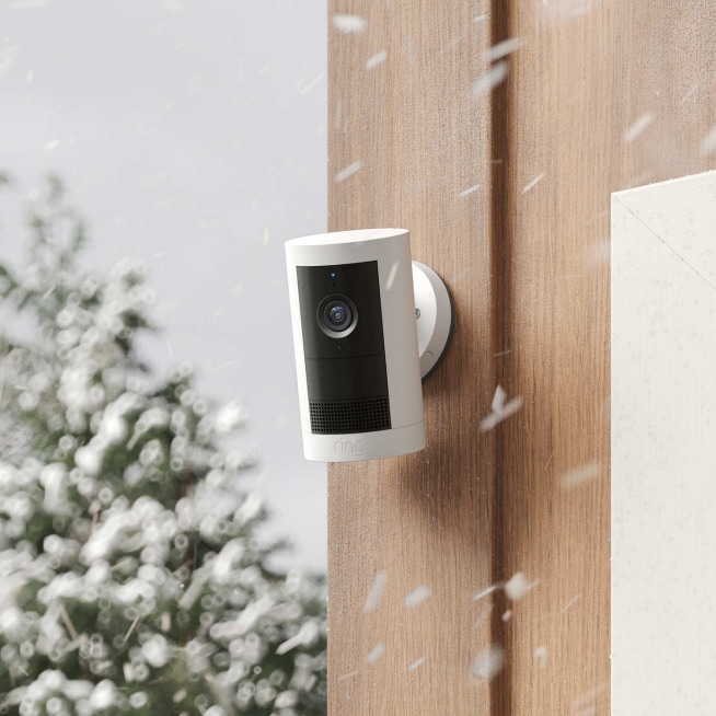 White Ring Outdoor Stick Up Cam mounted on a wooden wall during a snowfall.