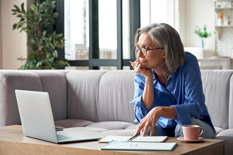 Woman with gray hair sitting on a couch with her laptop, documents, and coffee on the coffee table in front of her.