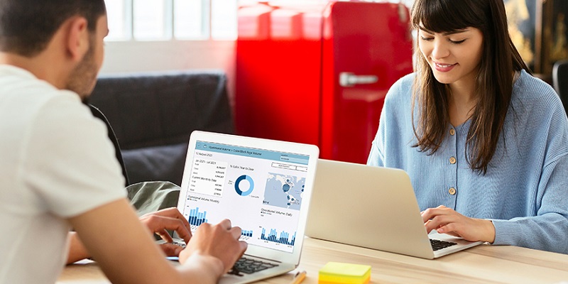 Man and woman sitting on opposite sites of a table using laptops. The man's laptop screen is visible showing charts and graphs.