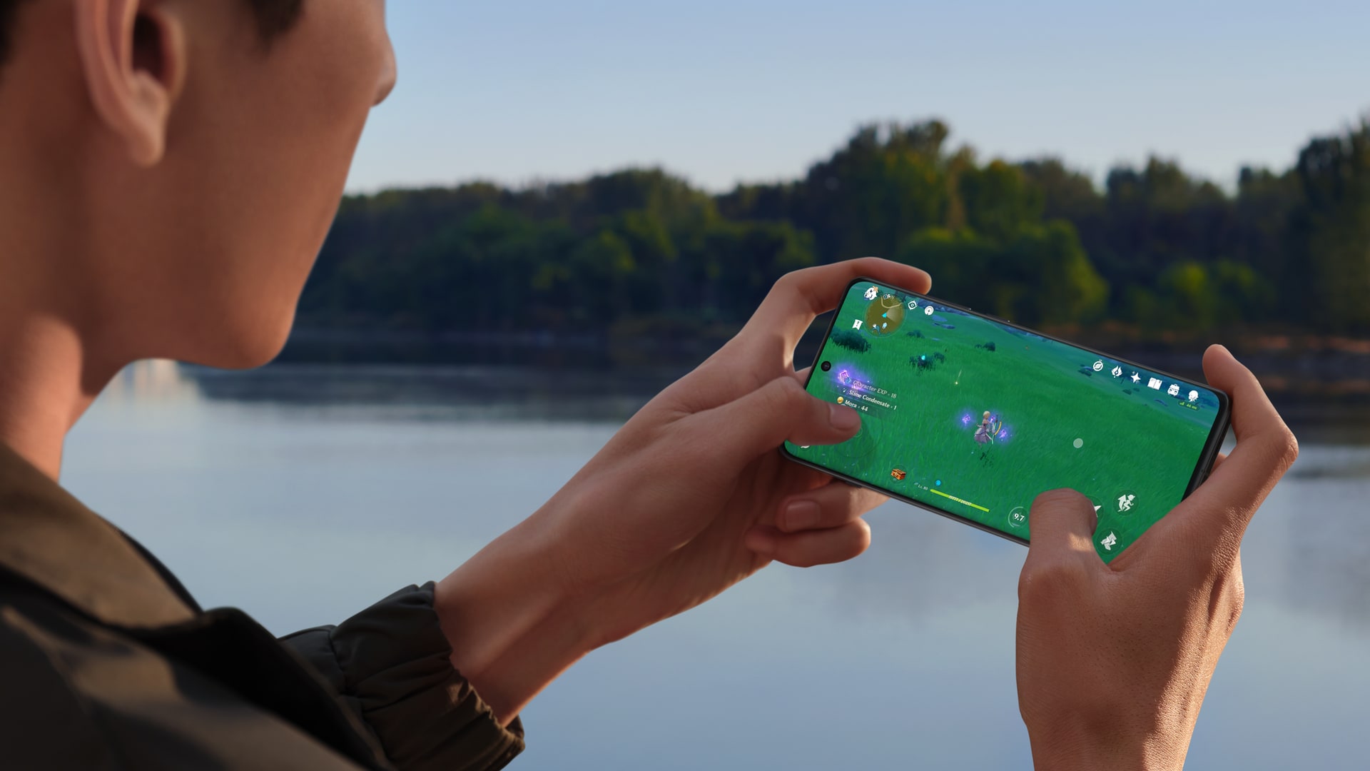 Ung man håller i en OnePlus 12 med båda händerna och spelar ett mobilspel, i bakgrunden en sjö eller ett vattendrag samt gröna träd