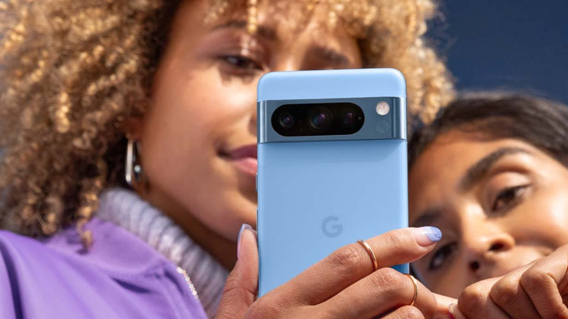 Två kvinnor tittar på en Google Pixel 8 Pro, blå mobiltelefon med metallband upptill som innehåller kameror och blixt, centrerad G-logo