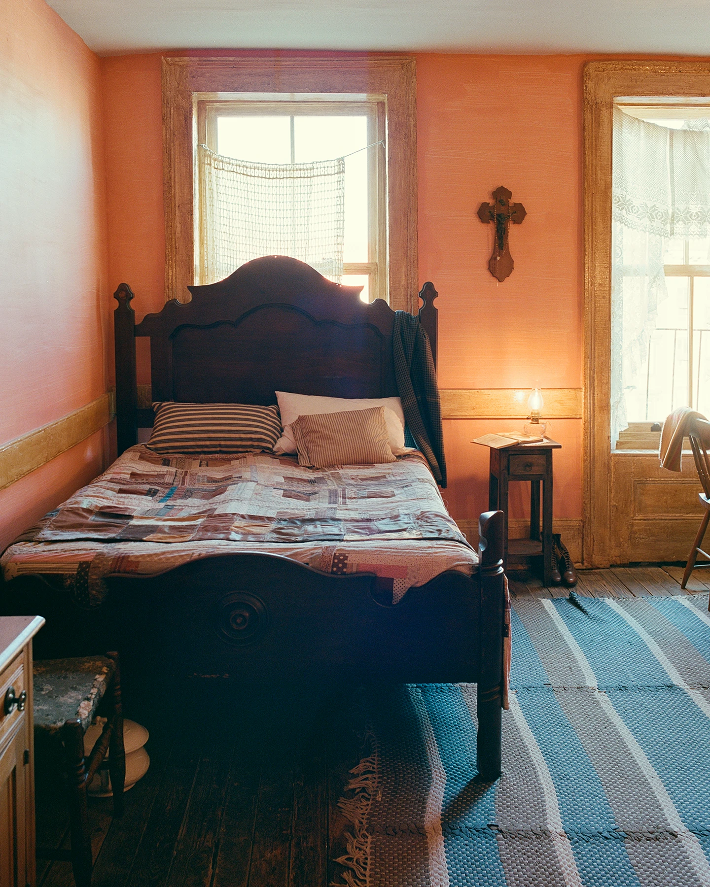 An ornamentally decorated wood bed holds a quilt and pillows. A lantern sits on a side table next to the bed and illuminates a wooden cross that hangs on the wall above it..