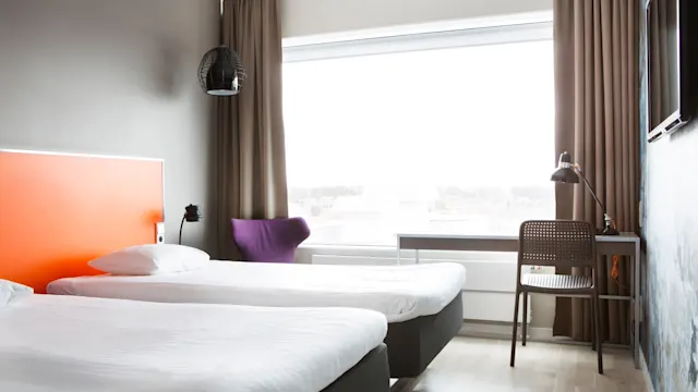 Två prydligt bäddade enkelsängar står i ett ljust hotellrum med ljust trägolv. En lila stol, skrivbord och lampa är nära ett stort fönster med bruna gardiner.