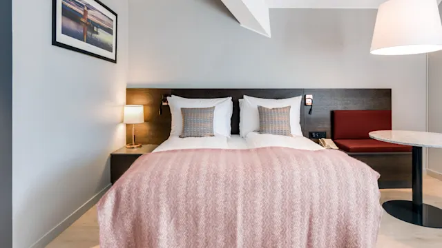 En pent redd dobbeltseng med mønstrede puter og et rosa teppe står i et moderne rom, ledsaget av en sidelys, veggkunst og et rundt bord under en hengende lampe.
