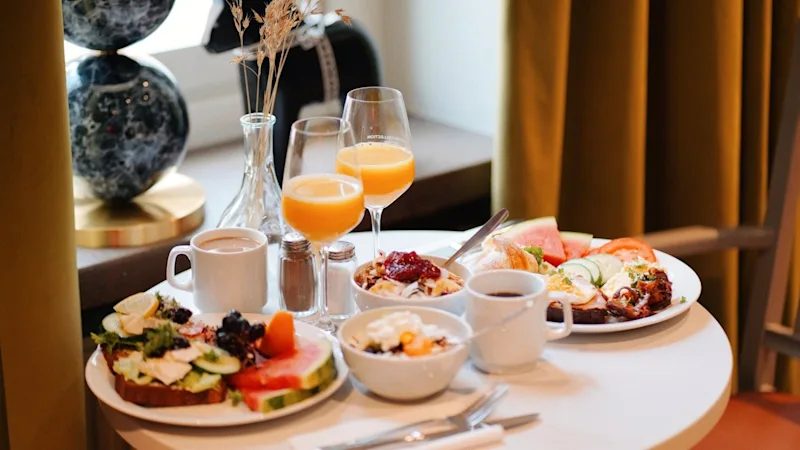 A breakfast table with plates of assorted fruits and pastries, glasses of orange juice, and cups of coffee in a cozy, softly lit dining room near curtained windows.