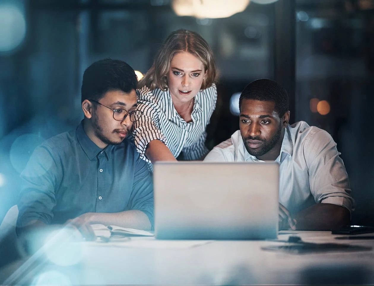 A woman stands between two men in front of a laptop and points to information on the screen