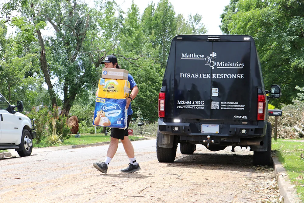 A man carries packages of paper towels and toilet paper from a black van on a neighborhood street.