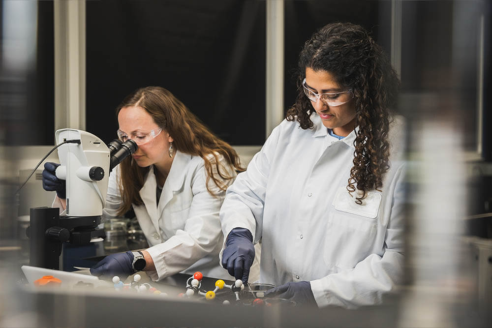 Two female scientists wear white lab coats, gloves, and safety goggles. One of the women is staring into a microscope.