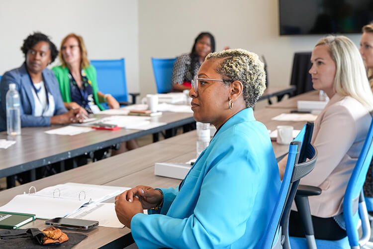 Six women of diverse backgrounds, dressed in business attire, sit around a conference table.