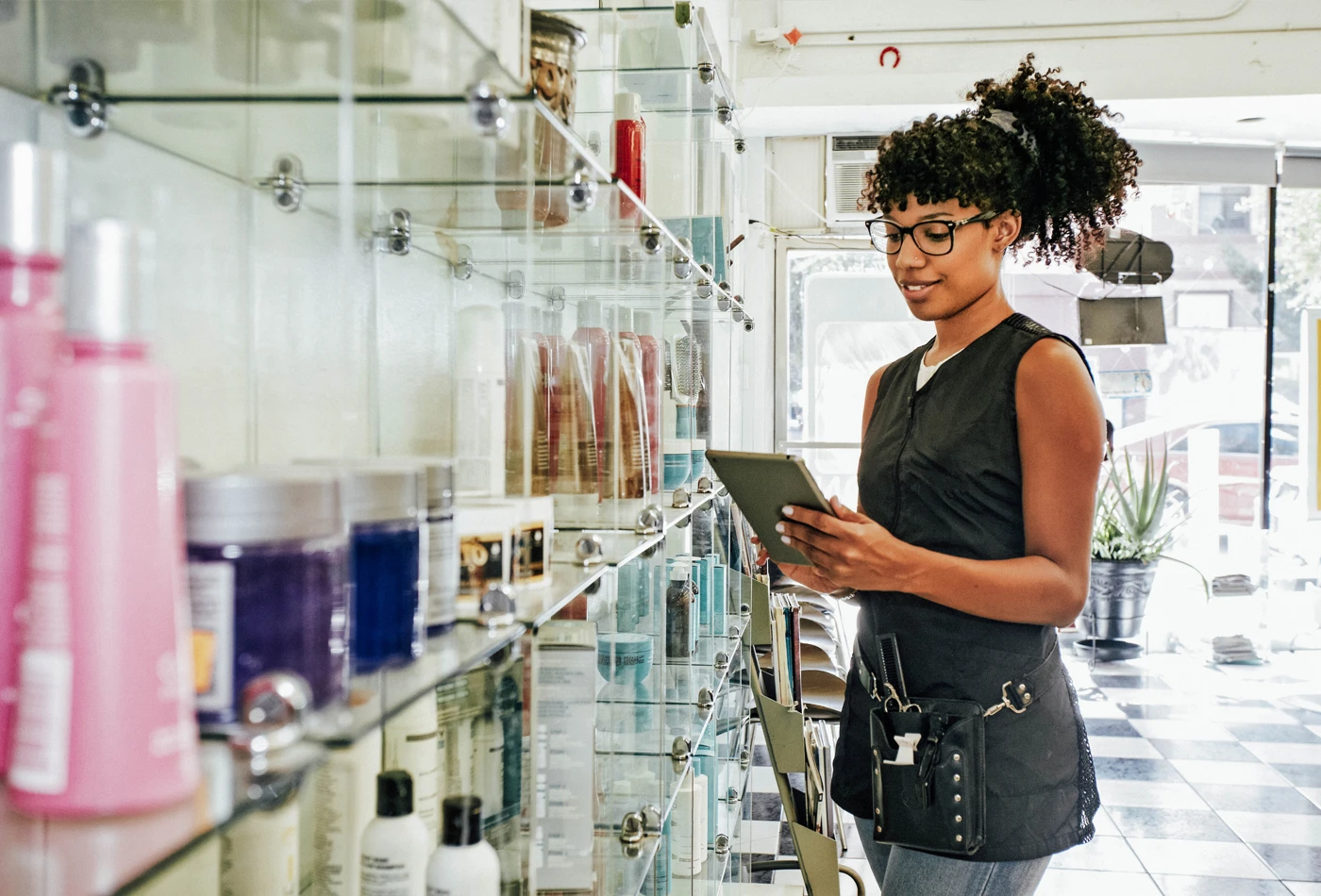 Woman with a tablet in hand at a salon taking inventory of products on a shelf in front of her.