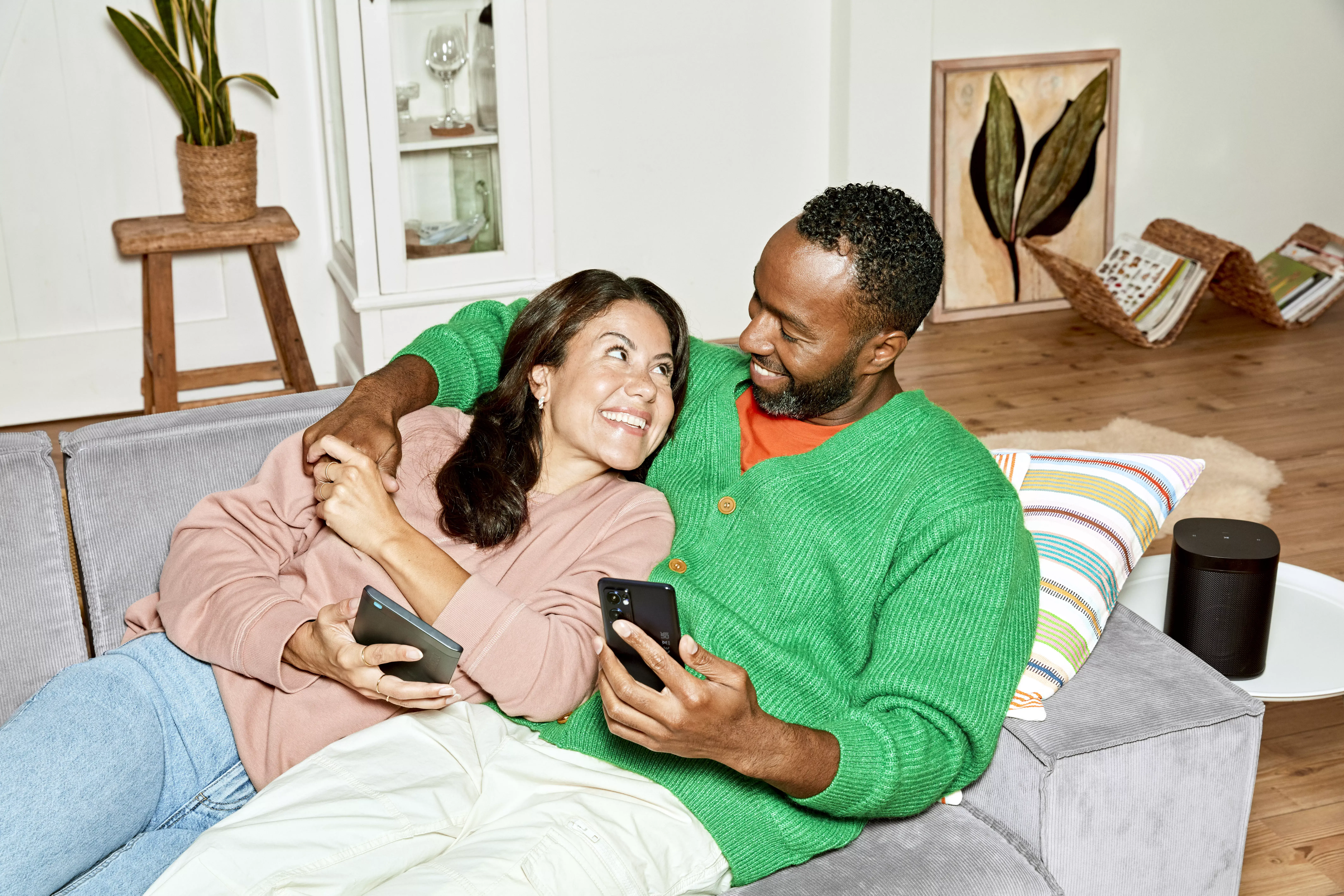 Een man en een vrouw liggen op de bank, houden elkaars hand vast en kijken elkaar blij aan. Ze hebben beide een mobiele telefoon in hun handen.