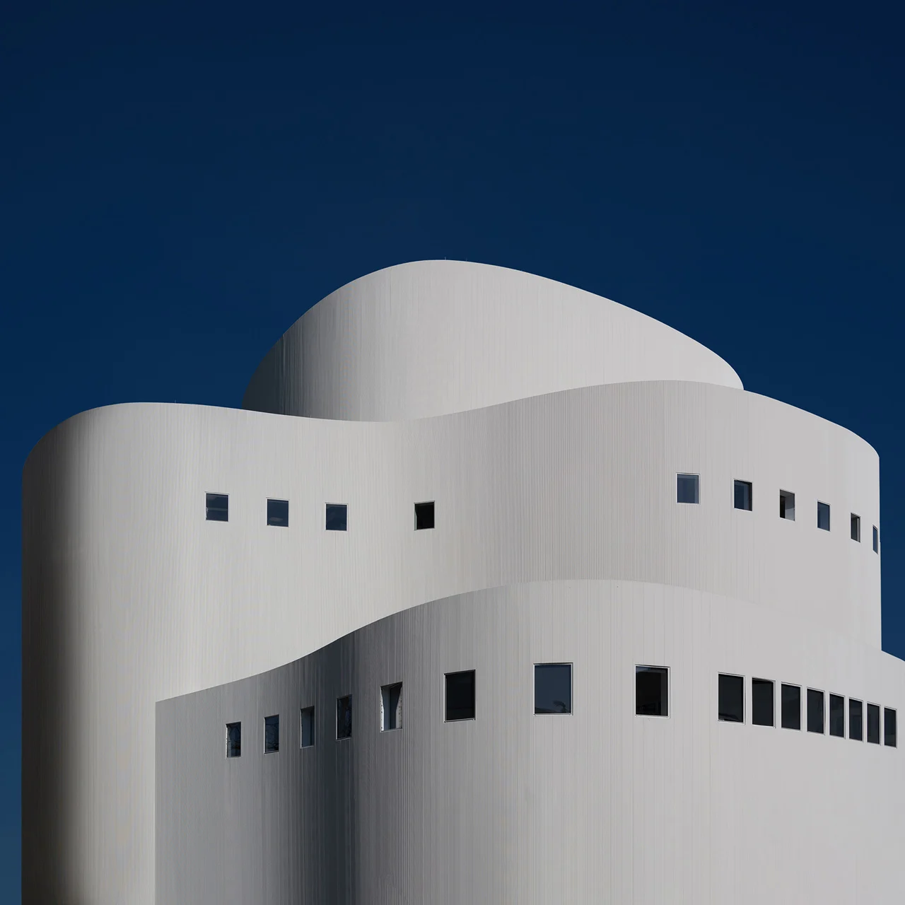 Edificio blanco con arquitectura curva y ventanas cuadradas contra un cielo azul.