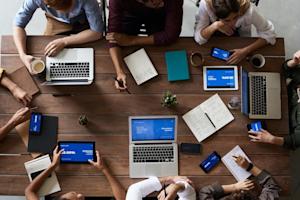 A group of employees sitting around a table and using several devices including laptops, tablets, and smartphones.