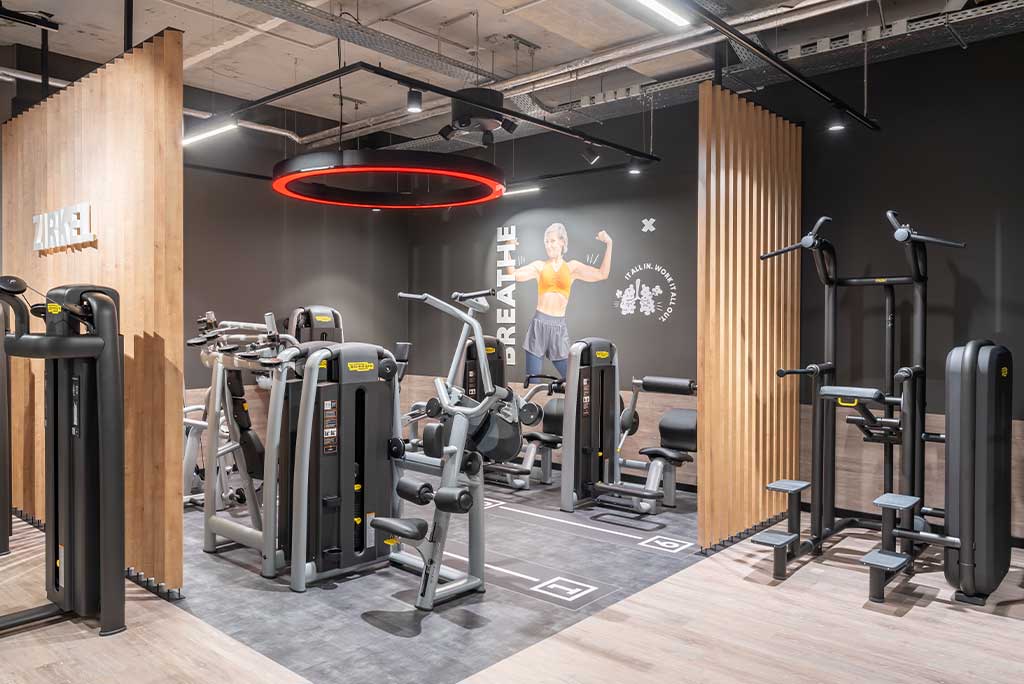 Zirkel-Bereich im FitX Fitnessstudio Berlin-Landsberger Allee, in dem verschiedene Trainingsgeräte kreisförmig angeordnet sind. Über den Geräten hängt ein großer Leuchtring, der in grün oder rot leuchtet und die Übungsdauer anzeigt.