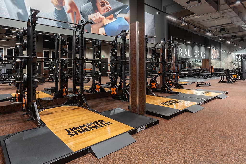 Weitläufiger Freihantelbereich mit verschiedenen Kraftgeräten im FitX Fitnessstudio Berlin-Waidmannslust. An der Seite sind Kurzhanteln vor einer Spiegelwand aufgereiht. An der oberen Wand prangt der Schriftzug „FOR ALL OF US“.