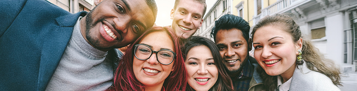 A group of six young adults of diverse backgrounds smiling and posing closely together for a selfie outdoors on a city street.