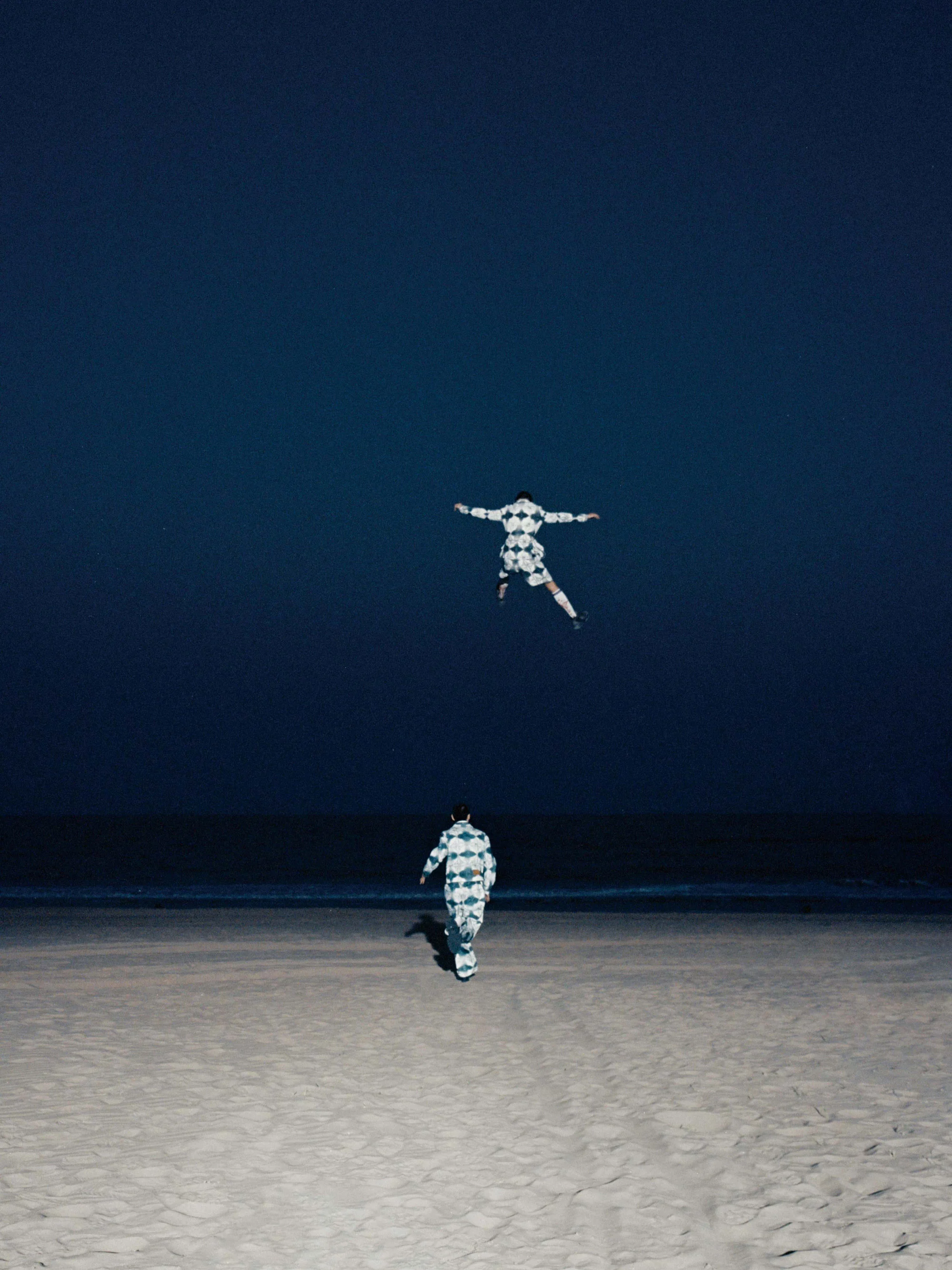 A photograph of models Jiao Tong and Wu Guoqiang facing away from the camera as they cross a sandy beach towards the ocean, one of them suspended in mid-air as if jumping with joy.