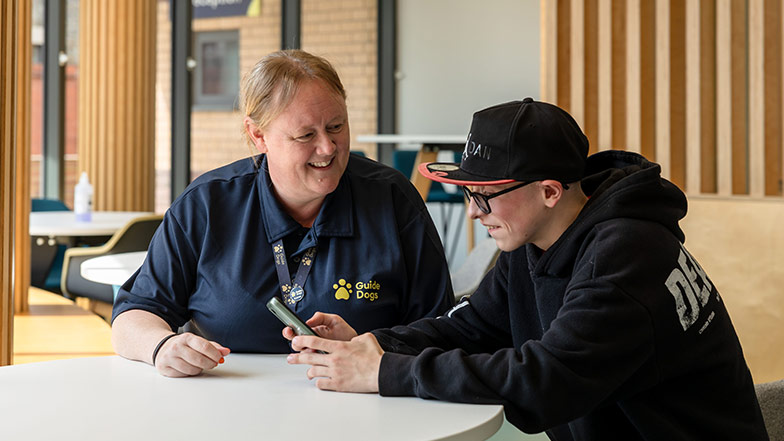 Kim, Habilitation Specialist, and Rhys, a service user, sit at a table together. They're both smiling, and Rhys is holding a smart phone.