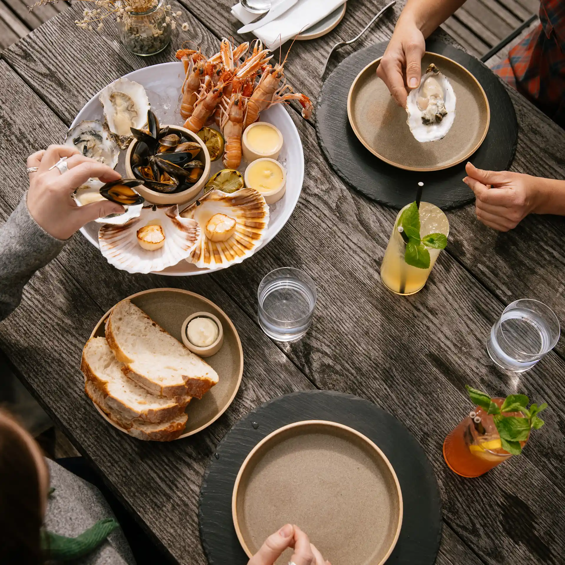 Van bovenaf zie je een houten tafel met twee mensen tegenover elkaar. Er staat een schaal met zeevruchten op tafel, samen met een bord brood en boter en verschillende cocktails.