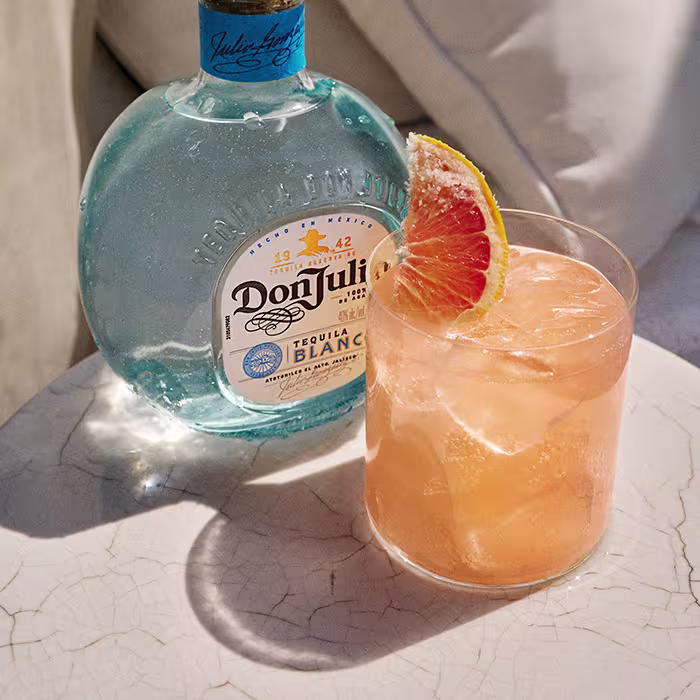 A bottle of Don Julio Blanco tequila on a table next to a Paloma cocktail with a grapefruit slice