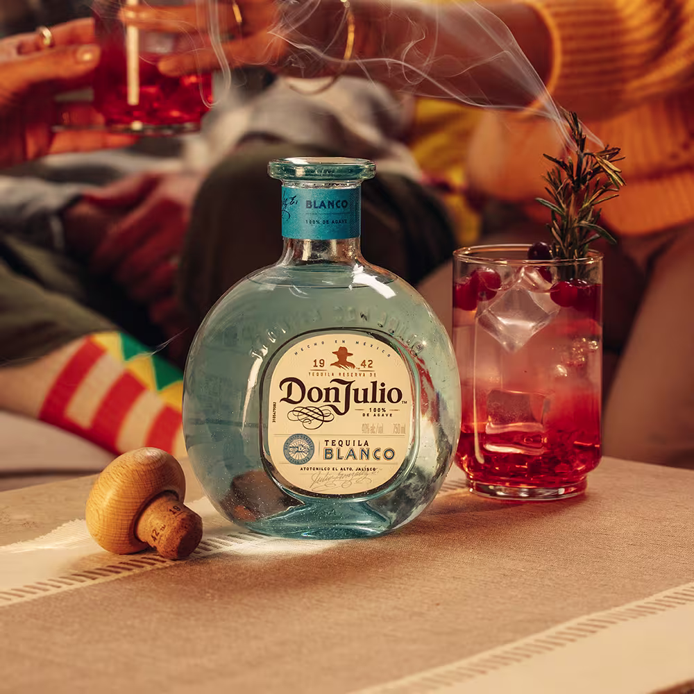 A bottle of Don Julio Blanco on a table next to a glass filled with Cranberry Rosemary Margarita cocktail