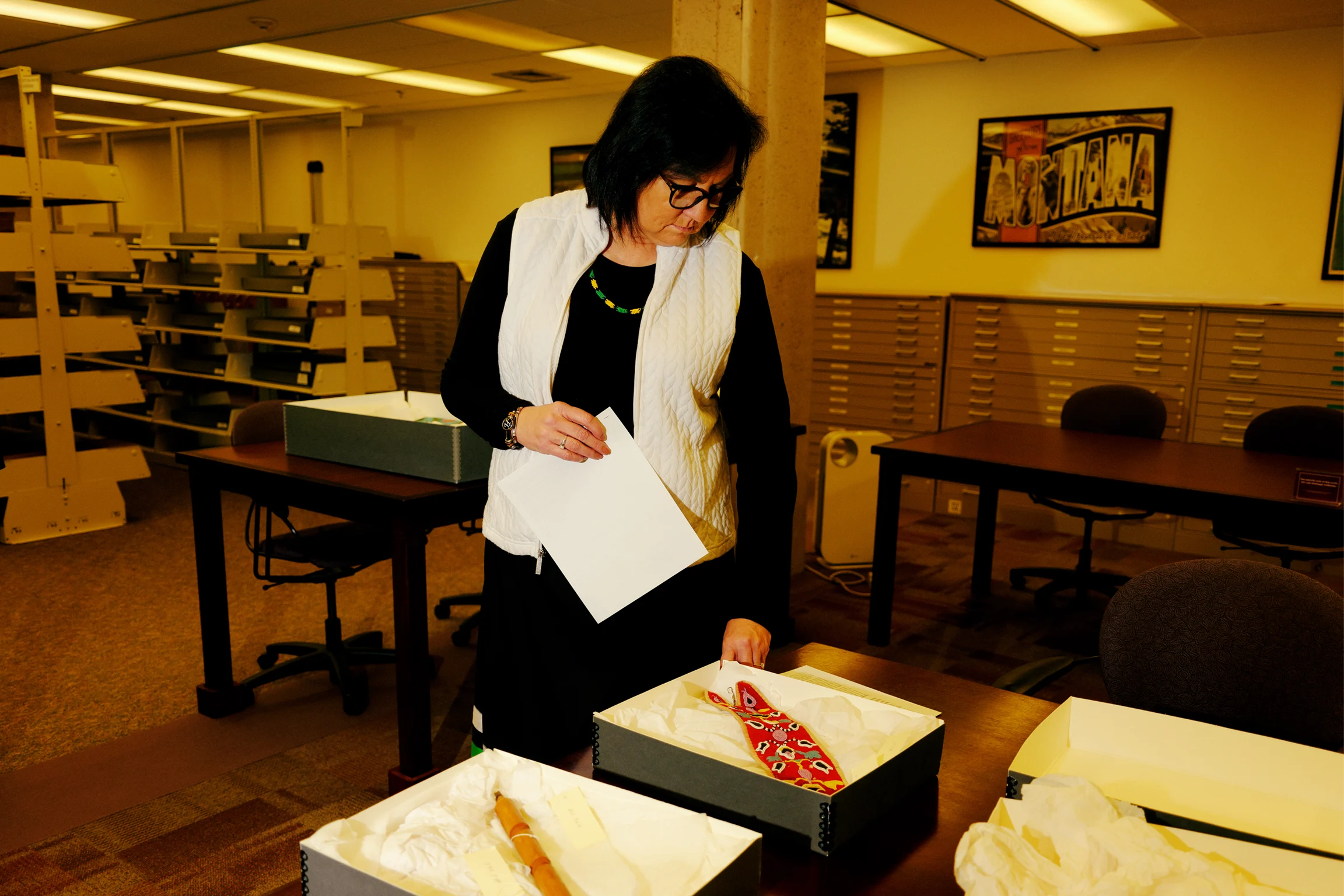 A Native woman with dark hair and glasses in a black long sleeve shirt and white vest stands in an archive room examining artifacts on a table