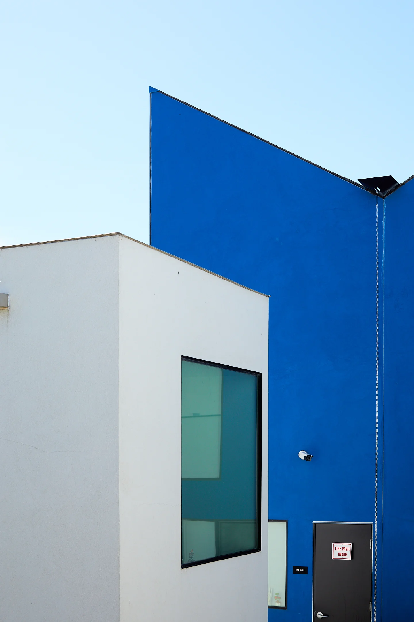 A low rectangular building with a window sits in front of a deep blue painted wall with a door and security camera on it