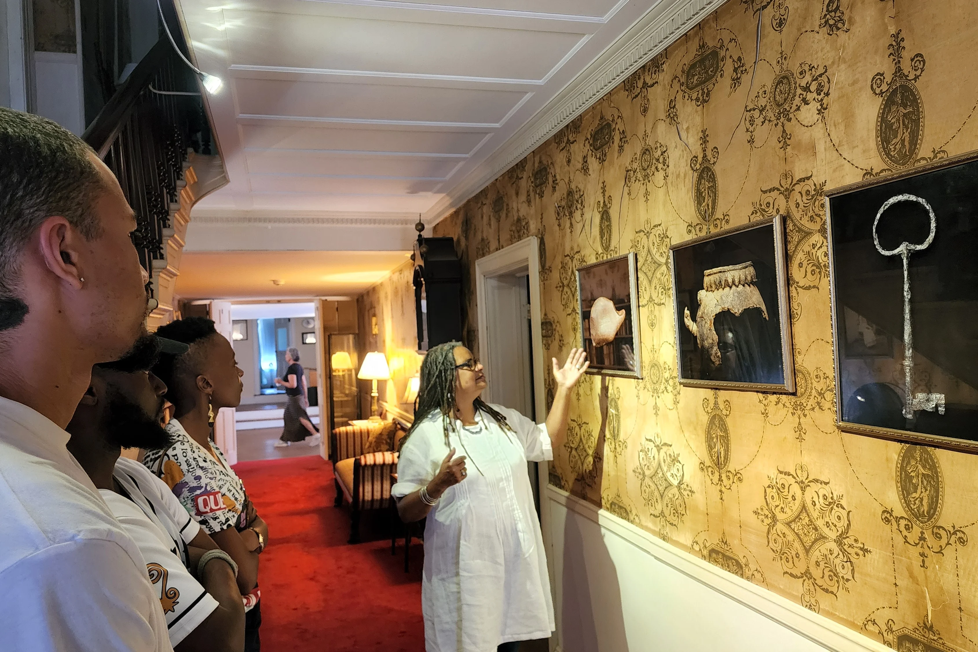 A historical home docent gestures to artworks hanging on a wall that is adorned with decorative wallpaper.