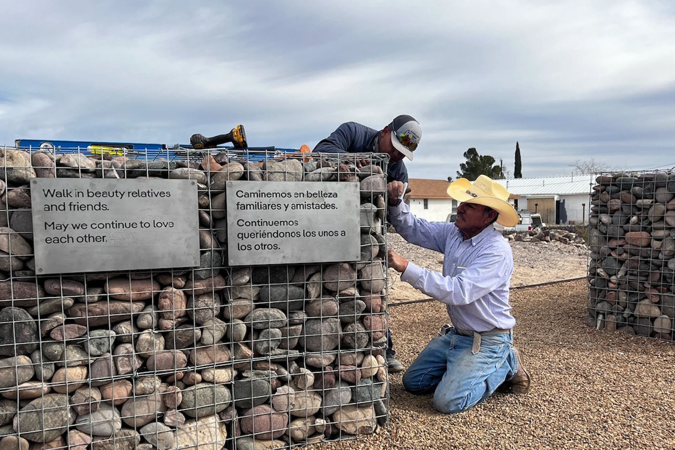 A man wearing a white cowboy hat and jeans kneeling on the ground beside a low stone wall