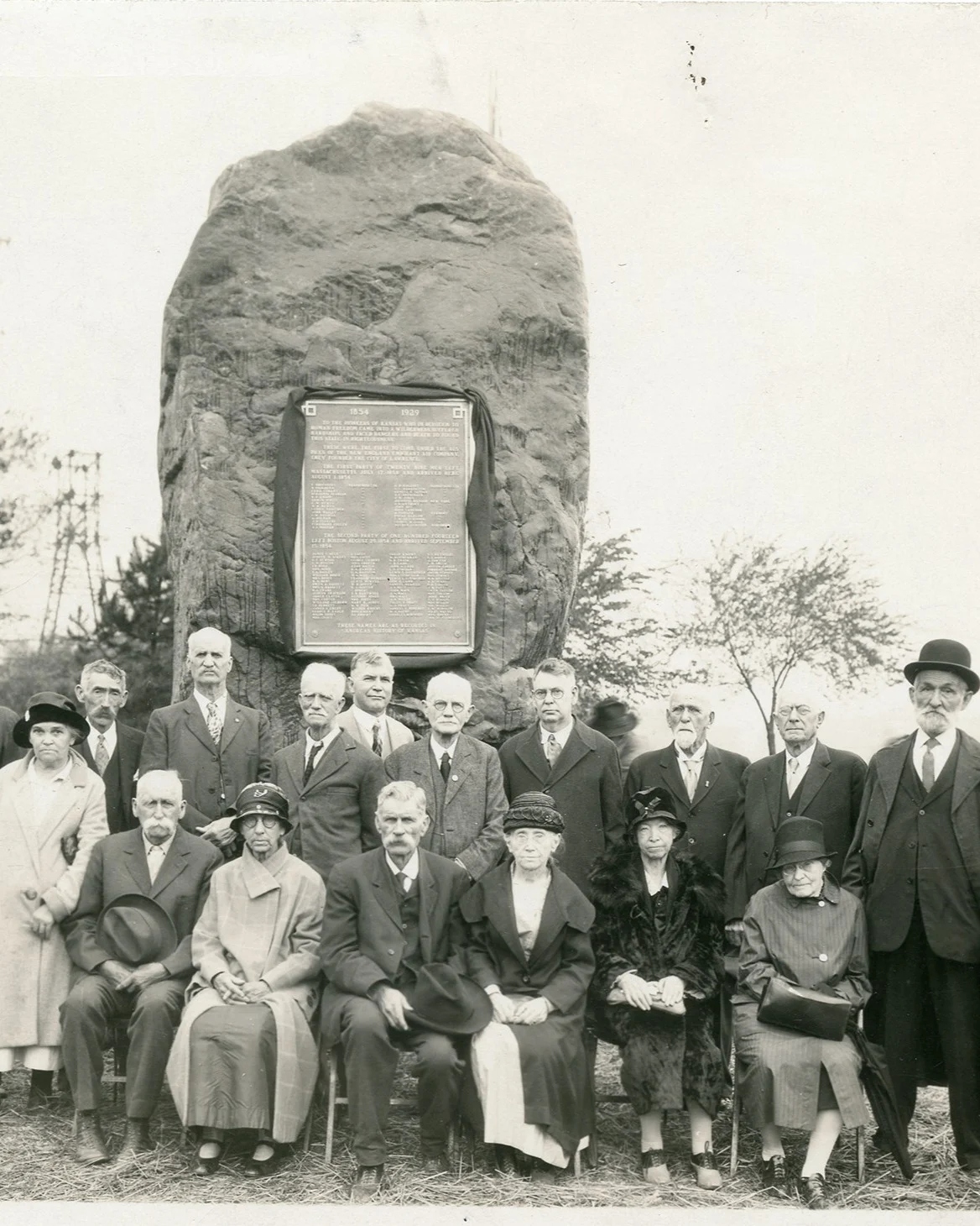 In sepia, sixteen white people gather in front of the Shunganunga boulder with a plaque affixed to it. The clothing pictured, suits and fur coats, appears to convey a high level of wealth.