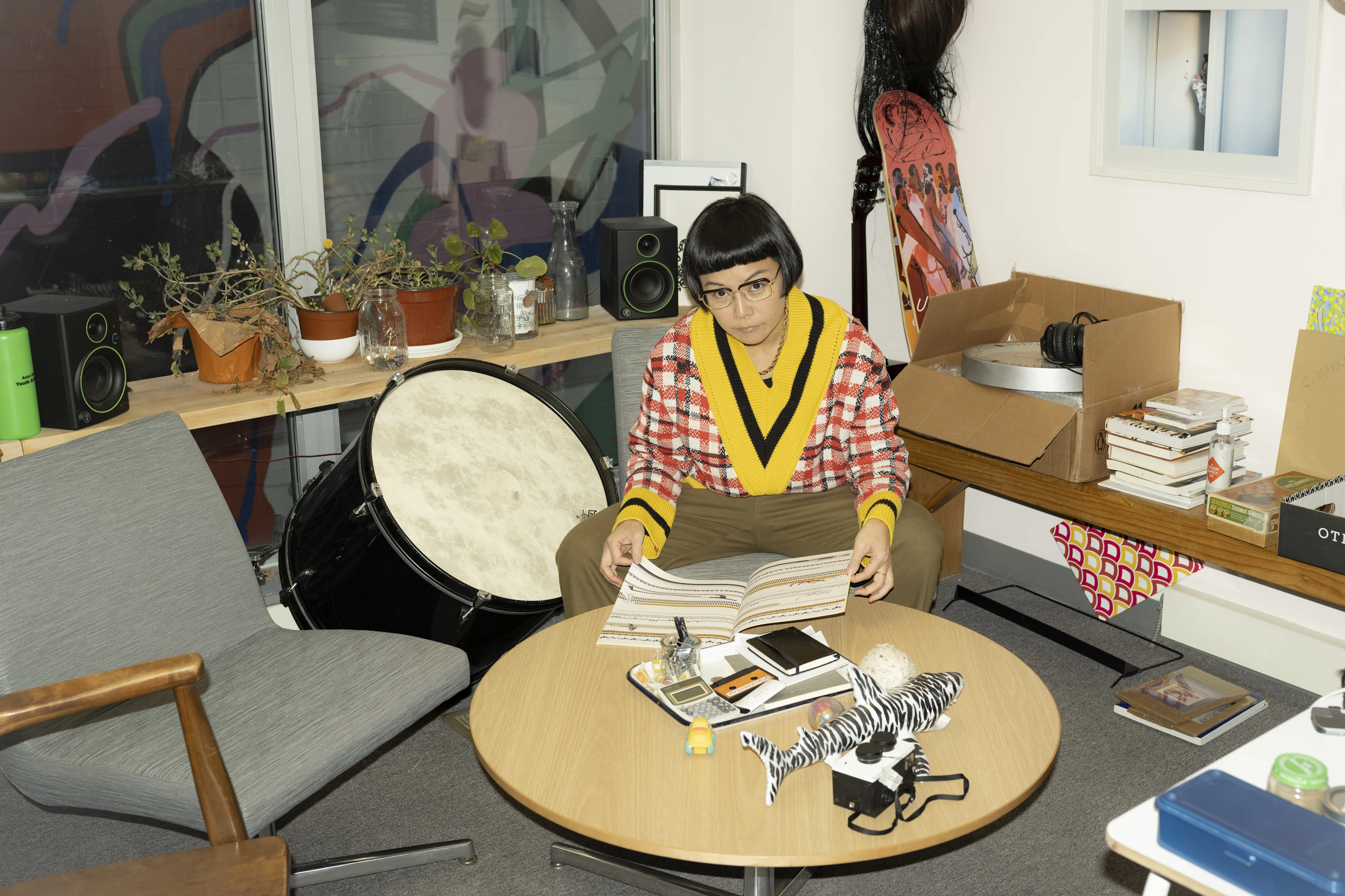 A person with short hair is sitting at a circular table. They are surrounded by assorted objects, books, and plants