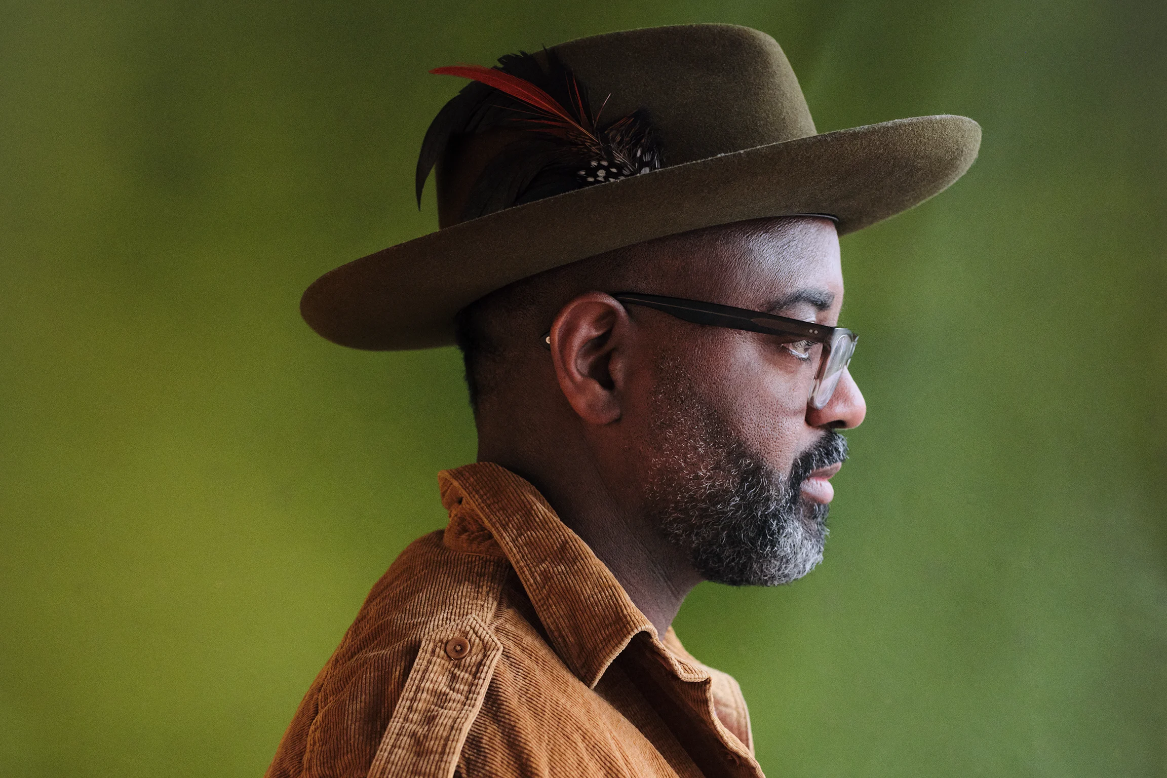 A side profile of a Black poet wearing glasses and a hat with a small feather hat pin.
