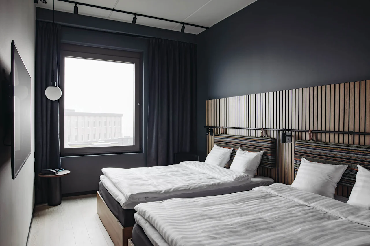 Two neatly made beds, topped with white linens, rest in a modern room. A large window reveals an urban building view, surrounded by dark walls and curtains.