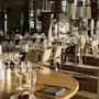 Et spisebord med pent arrangerte vinglass og bestikk, omgitt av trestoler. Mykt sollys filtrerer gjennom store vinduer, og gir en varm og innbydende atmosfære til restauranten.