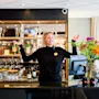 A smiling woman with open arms stands behind a hotel bar with glasses and flowers, welcoming guests.