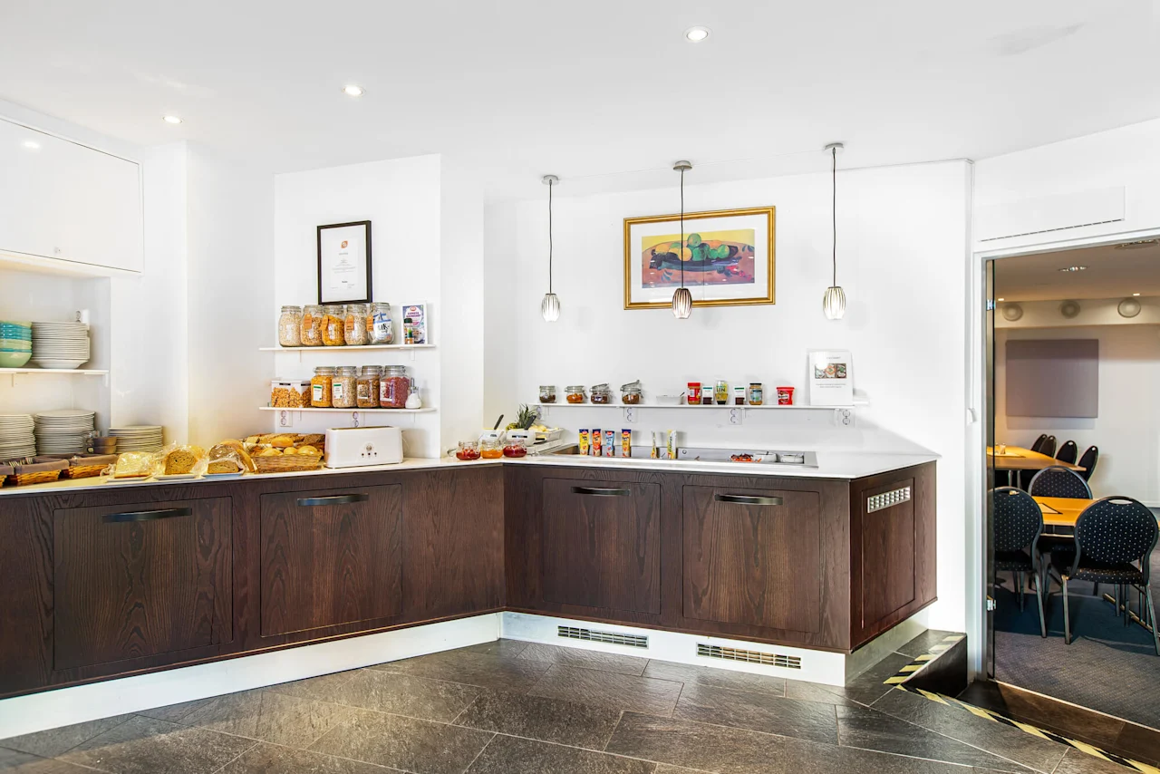 Breakfast buffet with jars of cereals, bread, and condiments on a wooden counter. Plates stack to the left. Three pendant lights hang above. A framed picture decorates the wall.