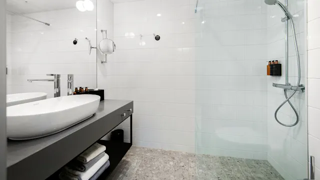 A modern bathroom features a double sink vanity with chrome faucets, a large wall mirror, and a glass-enclosed shower. The environment is minimalist with white tiles.