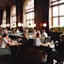 Folk spiser og snakker i en romslig, elegant designet restaurant med høye vinduer, moderne sirkulære lamper og frodig grønt på vinduskarmene. Atmosfæren er livlig og innbydende.