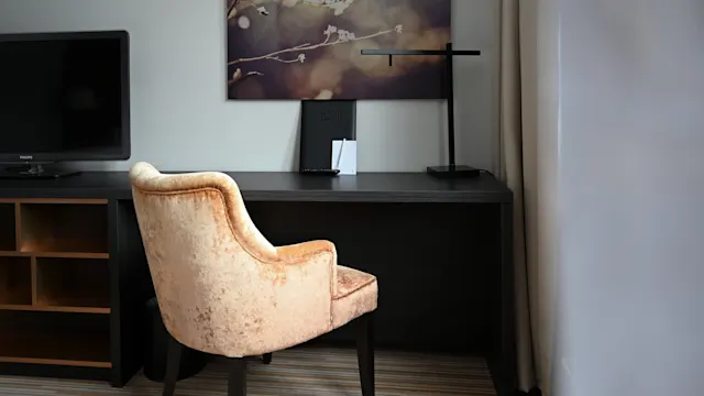 En beige stol står mot ett träskrivbord med en lampa och ett "WELCOME"-kort. En TV står bredvid skrivbordet, under en blomtema väggkonst.