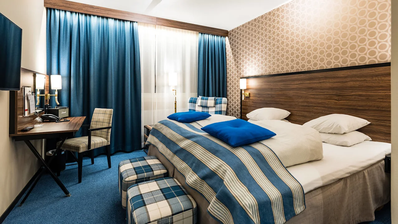 Et pent ordnet hotellrom med blå aksenter, mønstret tapet og oppredd seng som gir et inntrykk av gjestfrihet og komfort.