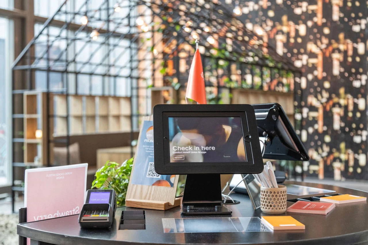 En digital incheckningsskärm visar "Check in here" på ett skrivbord med kortläsare, QR-koder och broschyrer. En metallstruktur och en vägg med växter syns i bakgrunden.