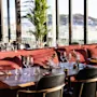 Tables set with wine glasses and cutlery rest against red cushioned seating in a sunlit restaurant with large windows and a view of city buildings and hills.
